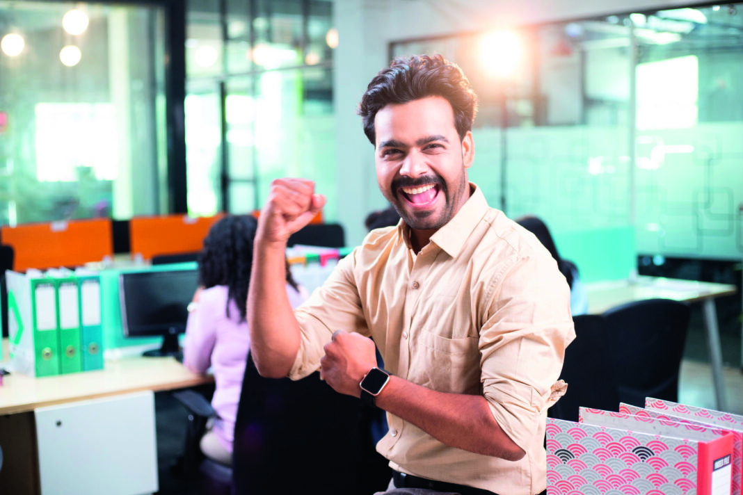 Portrait of happy young businessman with arms raised at office desk in office